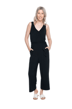 Petite model facing camera wearing black sleeveless, ankle-length, wide leg jumpsuit, featuring crossover neckline, waist belt and pockets. Jesse available in sizes 6-26