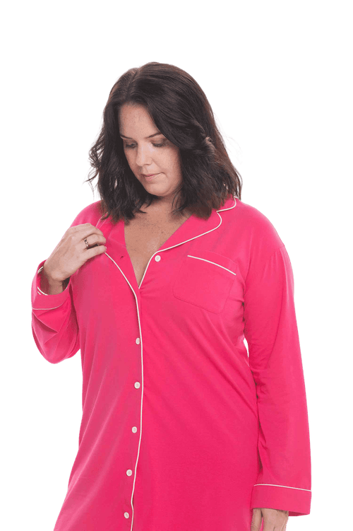 Brunette model facing camera wearing hot pink button up, mid thigh length pyjama shirt, featuring fold over collar, scooped hemline, and white piping. Parker available in sizes 6-18