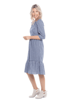 Model facing the side wearing blue gingham, mid-length sleeved midi dress, features rounded neckline, tiered skirt and pockets. Dorothy available in sizes 6-18