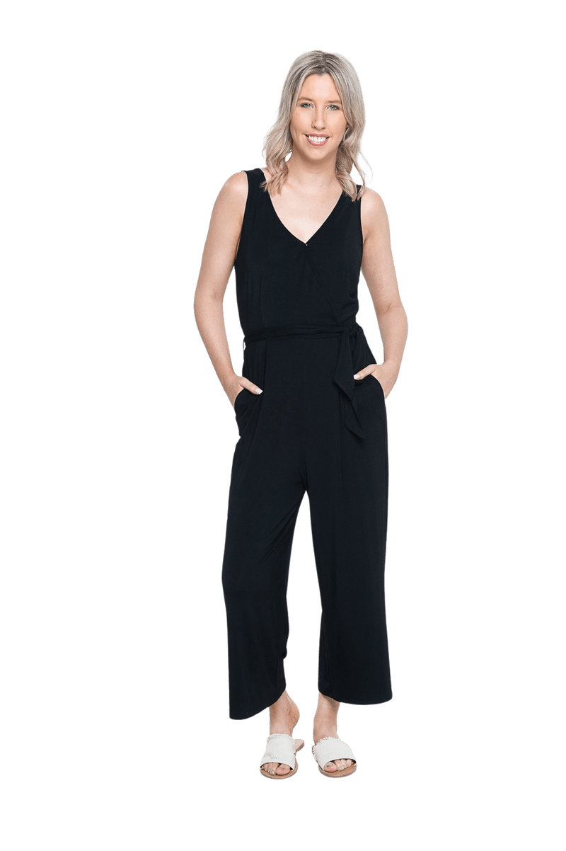 Petite model facing camera wearing black sleeveless, ankle-length, wide leg jumpsuit, featuring crossover neckline, waist belt and pockets. Jesse available in sizes 6-26