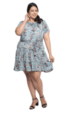 Curvy model facing camera wearing dusty blue with small pink floral, knee-length dress, featuring pockets, a rounded, frilled neckline, and a tiered skirt. Kirsty available in sizes 6-26