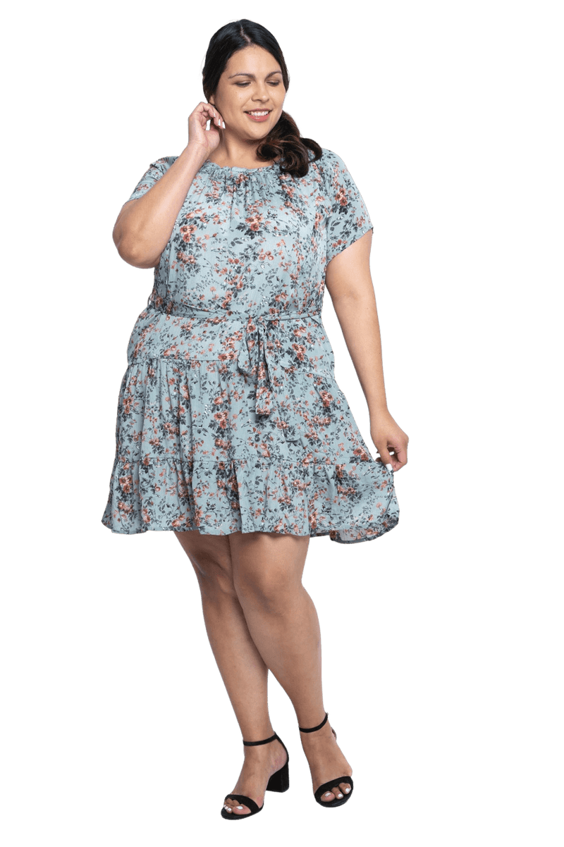 Curvy model facing camera wearing dusty blue with small pink floral, knee-length dress, featuring pockets, a rounded, frilled neckline, and a tiered skirt. Kirsty available in sizes 6-26