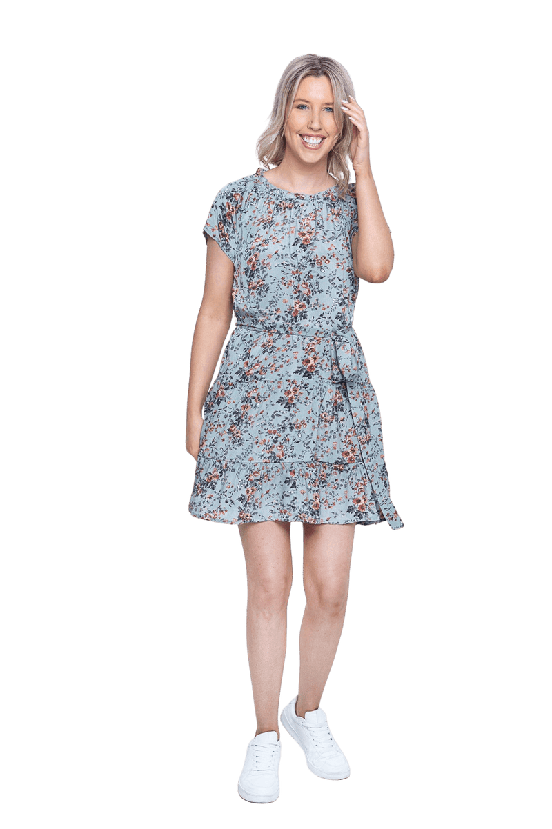 Petite model facing camera wearing dusty blue with small pink floral, knee-length dress, featuring pockets, a rounded, frilled neckline, and a tiered skirt. Kirsty available in sizes 6-26