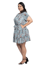 Curvy model facing the side wearing dusty blue with small pink floral, knee-length dress, featuring pockets, a rounded, frilled neckline, and a tiered skirt. Kirsty available in sizes 6-26