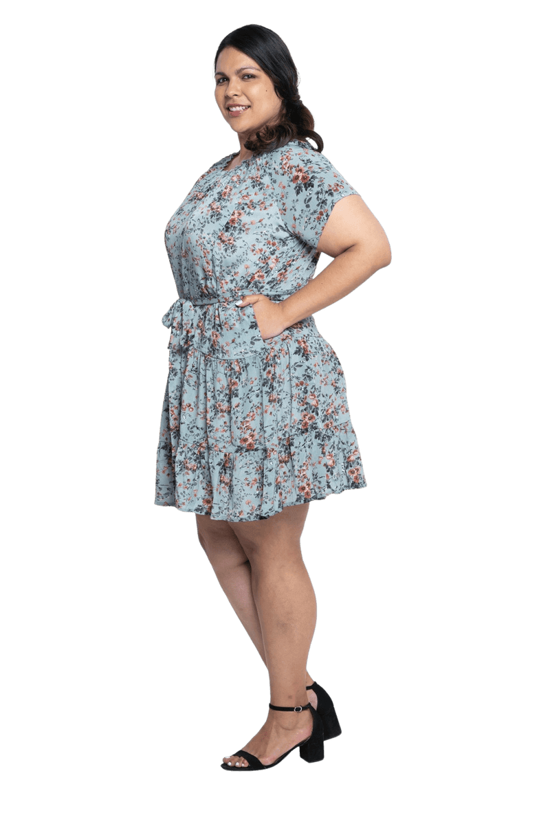 Curvy model facing the side wearing dusty blue with small pink floral, knee-length dress, featuring pockets, a rounded, frilled neckline, and a tiered skirt. Kirsty available in sizes 6-26