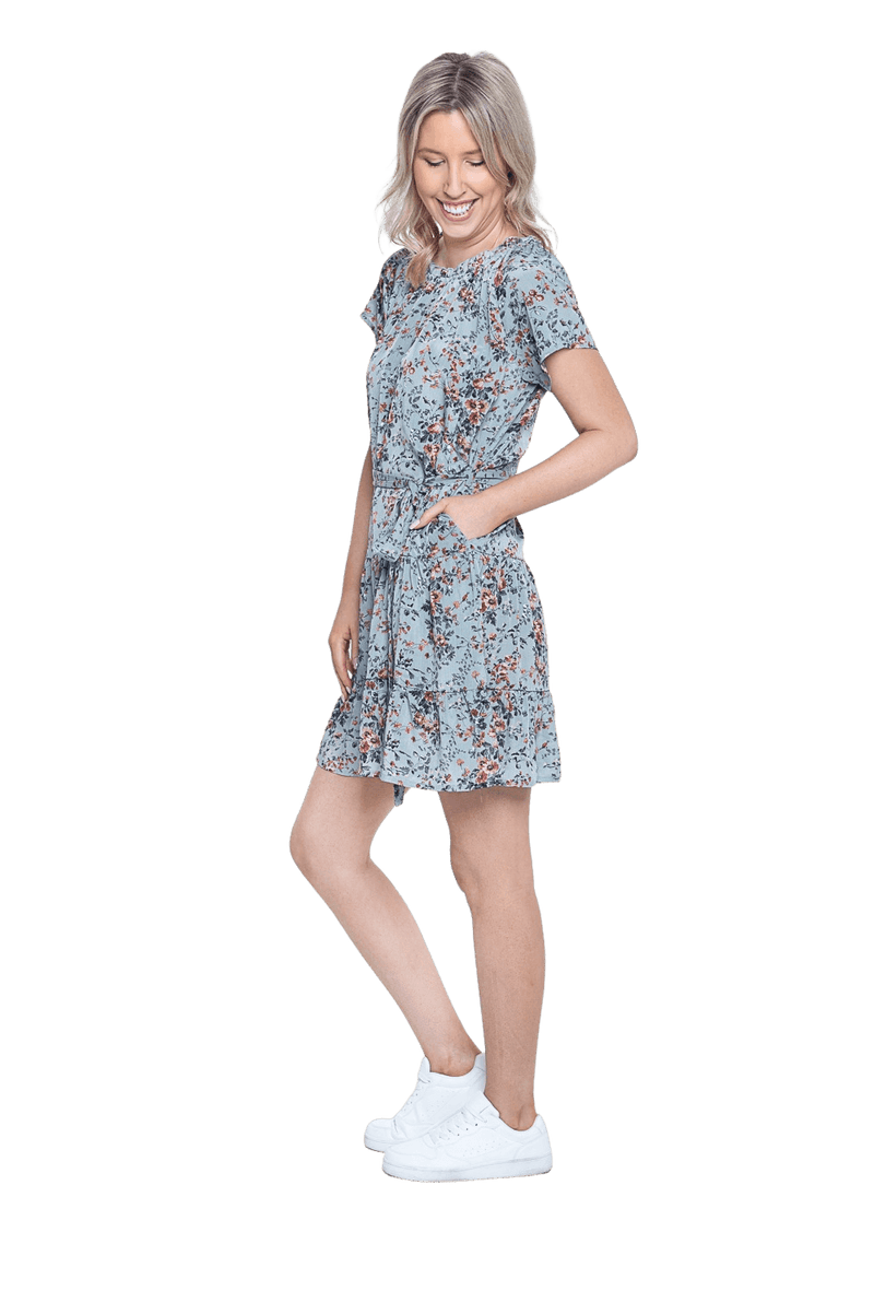 Petite model facing the side wearing dusty blue with small pink floral, knee-length dress, featuring pockets, a rounded, frilled neckline, and a tiered skirt. Kirsty available in sizes 6-26