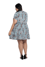 Curvy model facing the back wearing dusty blue with small pink floral, knee-length dress, featuring pockets, a rounded, frilled neckline, and a tiered skirt. Kirsty available in sizes 6-26