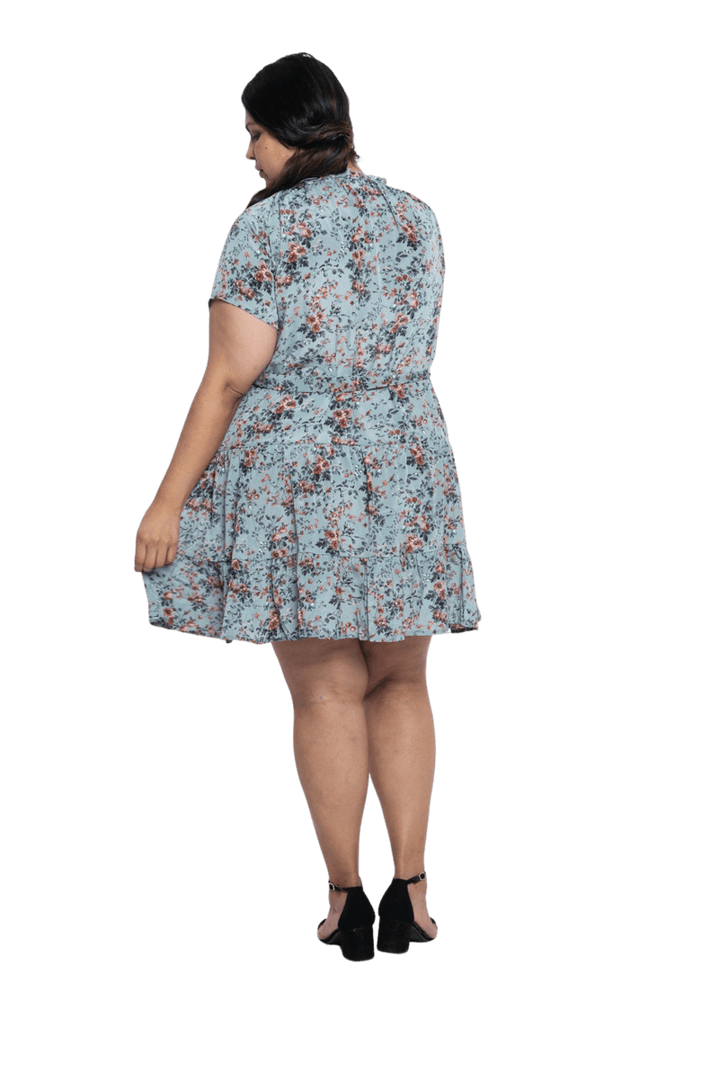Curvy model facing the back wearing dusty blue with small pink floral, knee-length dress, featuring pockets, a rounded, frilled neckline, and a tiered skirt. Kirsty available in sizes 6-26