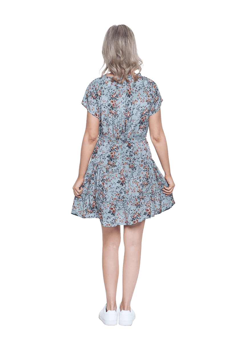 Petite model facing the back wearing dusty blue with small pink floral, knee-length dress, featuring pockets, a rounded, frilled neckline, and a tiered skirt. Kirsty available in sizes 6-26