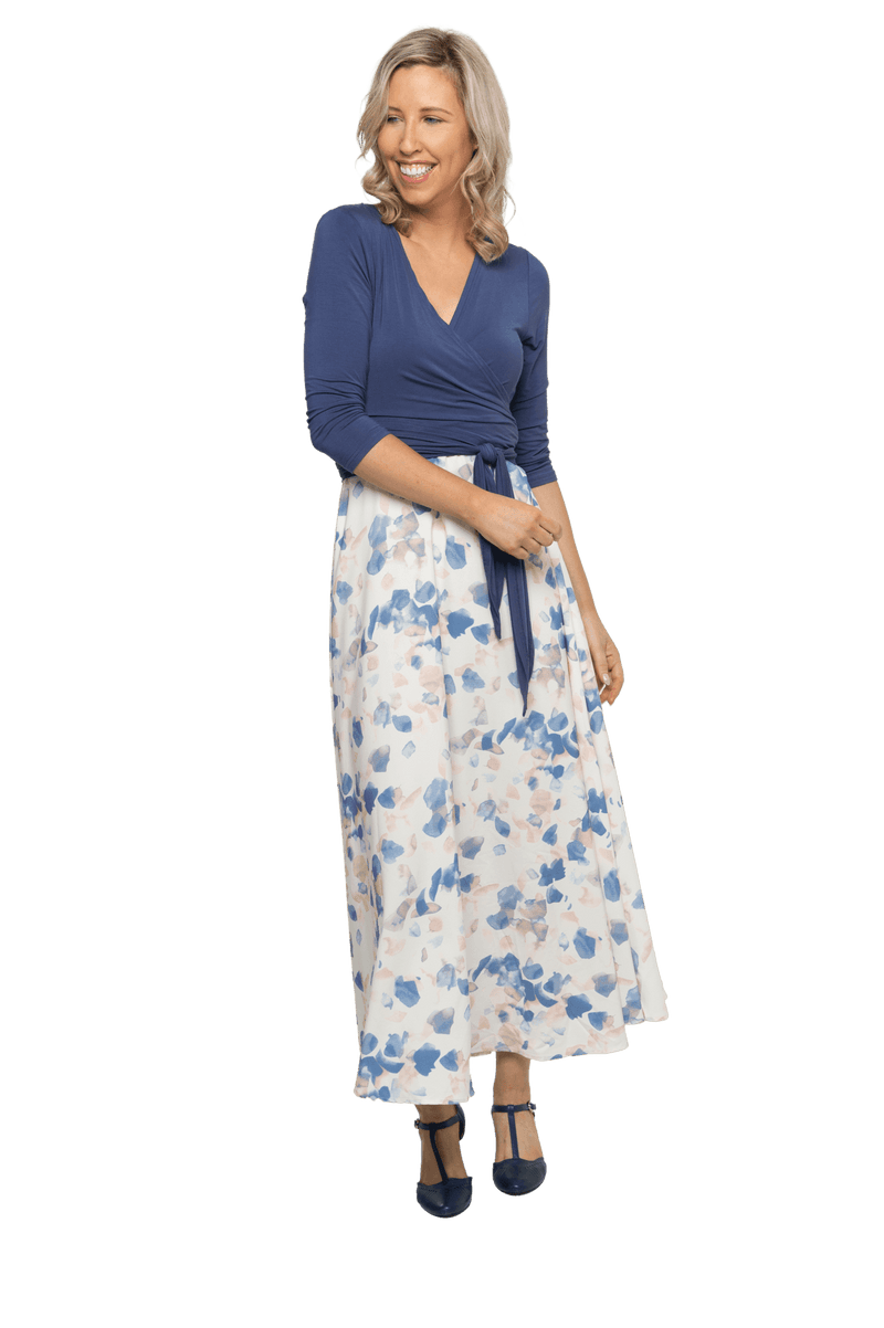 Petite model facing camera wearing maxi dress with blue mid sleeved crossover top attached to white skirt with blue and tan watercolour accents. Mai available in sizes 6-26