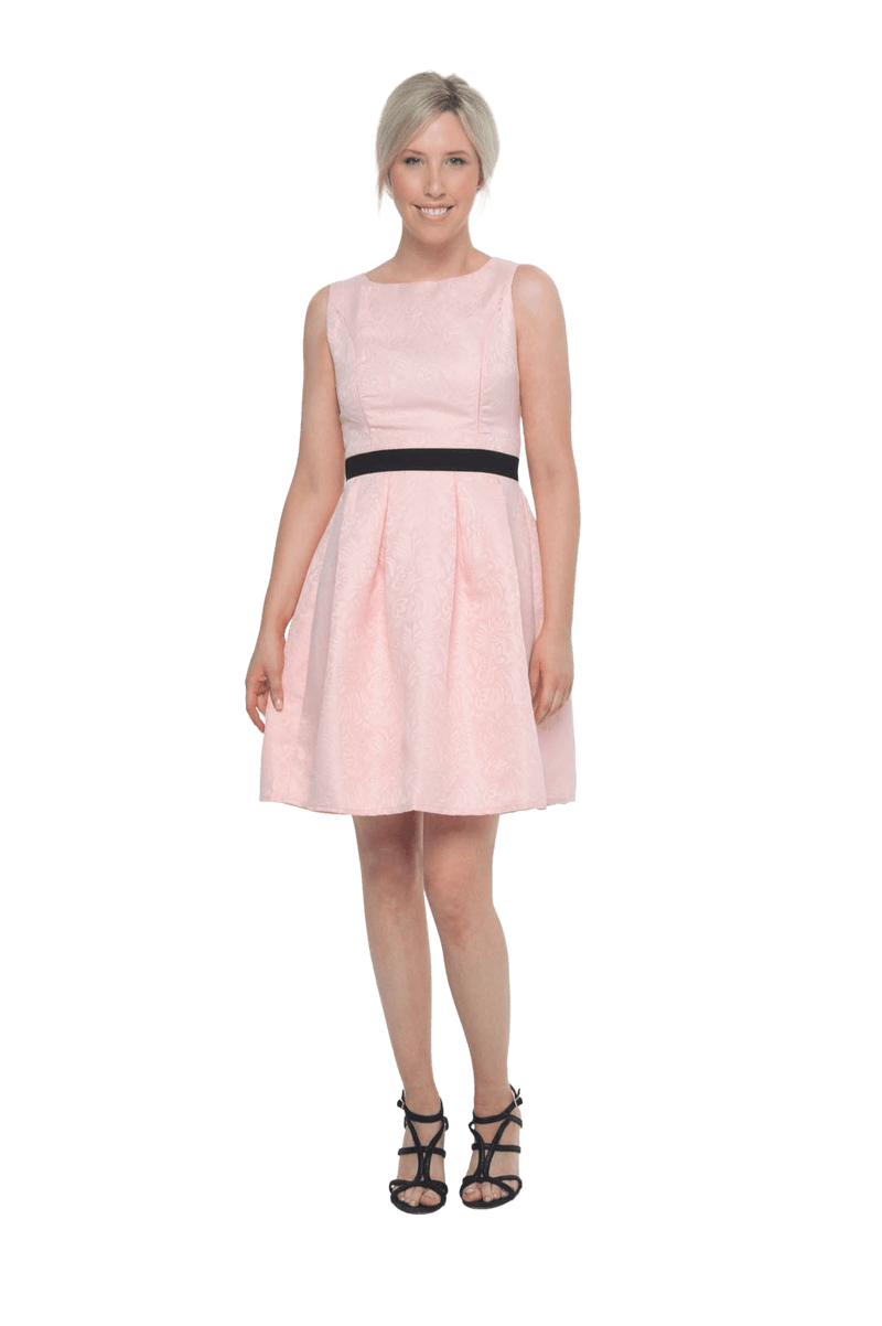 Petite model facing the camera wearing pink cocktail dress, featuring a boat neckline, back zip and a feature black sash across the waist. Marie available in sizes 6-18