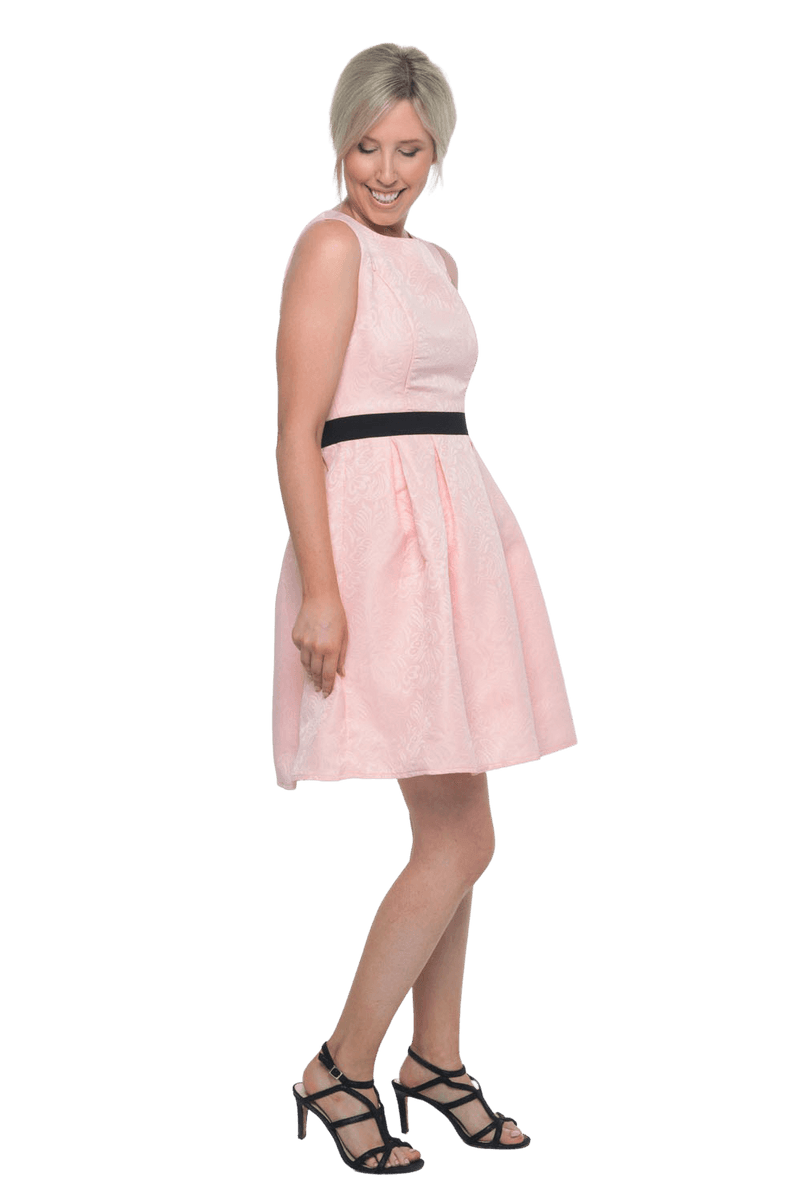 Petite model facing the side wearing pink cocktail dress, featuring a boat neckline, back zip and a feature black sash across the waist. Marie available in sizes 6-18