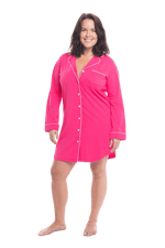 Brunette model facing camera wearing hot pink button up, mid thigh length pyjama shirt, featuring fold over collar, scooped hemline, and white piping. Parker available in sizes 6-18