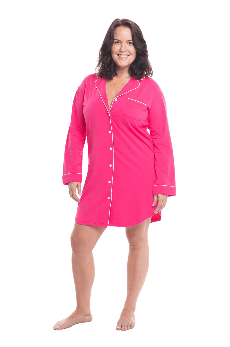 Brunette model facing camera wearing hot pink button up, mid thigh length pyjama shirt, featuring fold over collar, scooped hemline, and white piping. Parker available in sizes 6-18