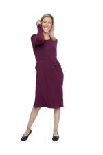 Petite model facing camera wearing long sleeved, knee length burgundy dress. Featuring pockets, a rounded neckline, and pleated front. Robyn available in sizes 6-26