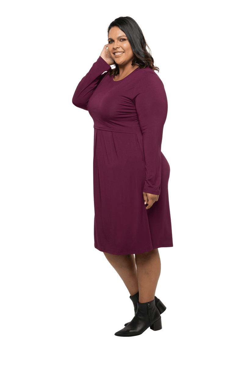 Curvy model facing the side wearing long sleeved, knee length burgundy dress. Featuring pockets, a rounded neckline, and pleated front. Robyn available in sizes 6-26