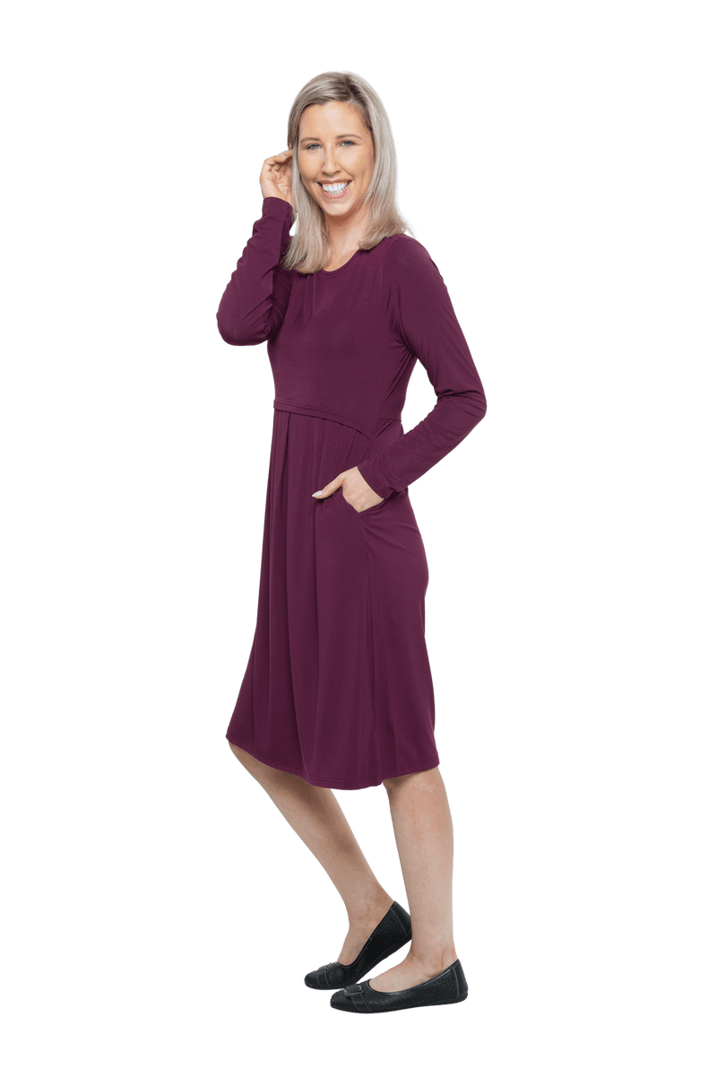 Petite model facing the side wearing long sleeved, knee length burgundy dress. Featuring pockets, a rounded neckline, and pleated front. Robyn available in sizes 6-26