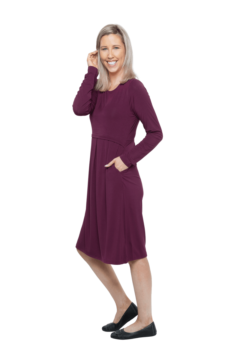 Petite model facing the side wearing long sleeved, knee length burgundy dress. Featuring pockets, a rounded neckline, and pleated front. Robyn available in sizes 6-26