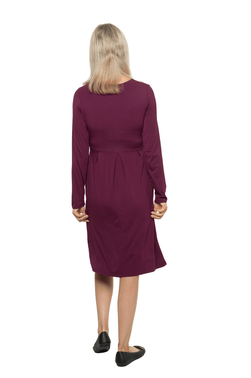 Petite model facing the back wearing long sleeved, knee length burgundy dress. Featuring pockets, a rounded neckline, and pleated front. Robyn available in sizes 6-26